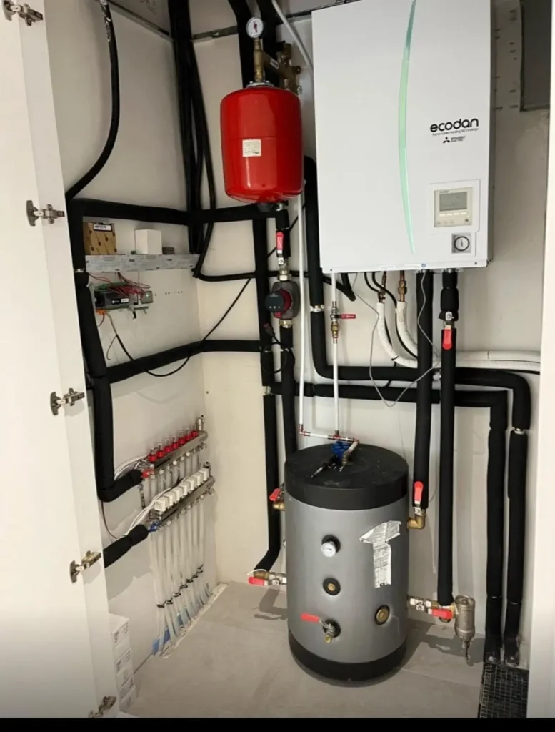 Instalación técnica de aerotermia con unidad Ecodan, depósito acumulador, vaso de expansión y colectores en un armario técnico.