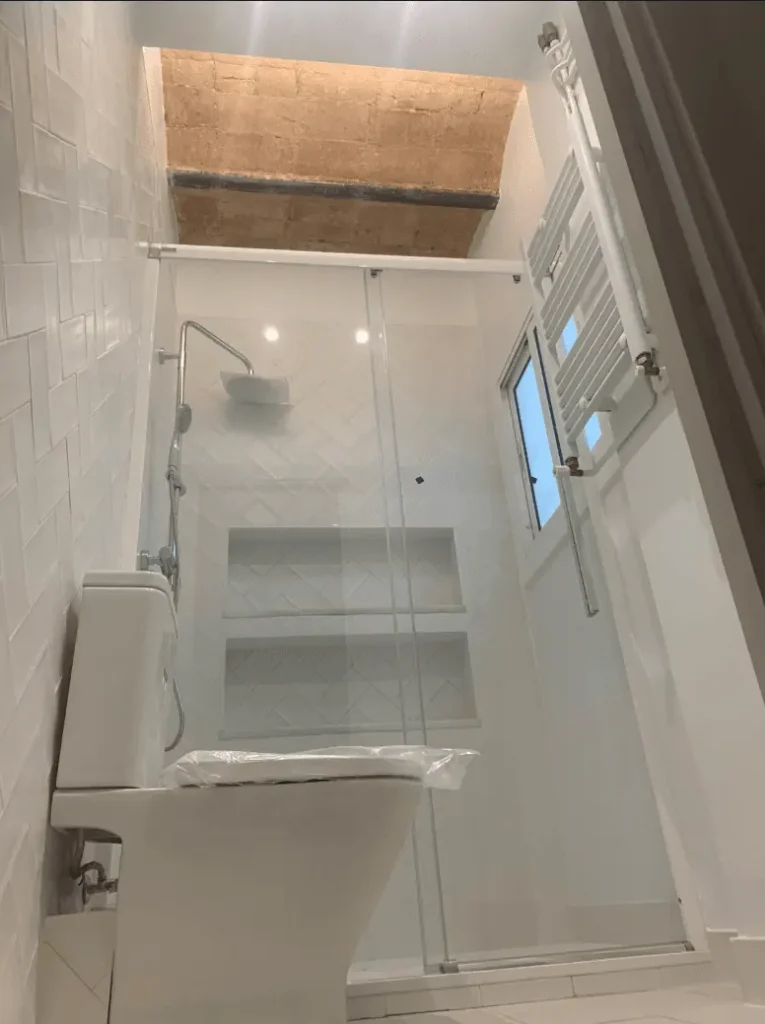 Baño moderno con ducha acristalada, estanterías empotradas y techo rústico con vigas de madera.