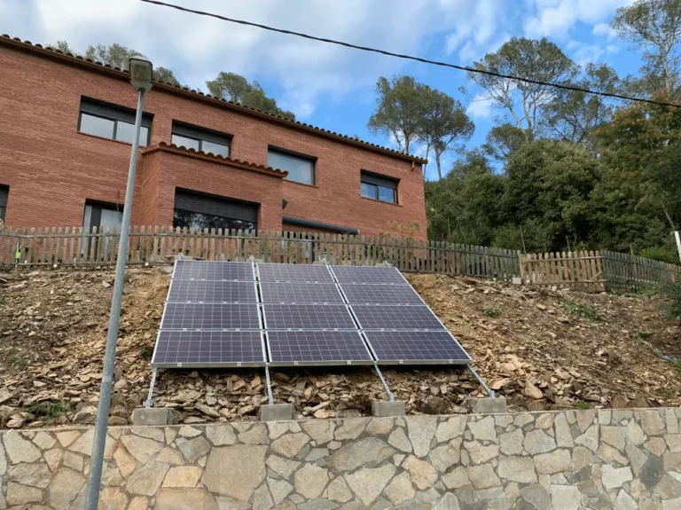 Paneles solares fotovoltaicos instalados en un terreno inclinado frente a una casa de obra vista en Girona.