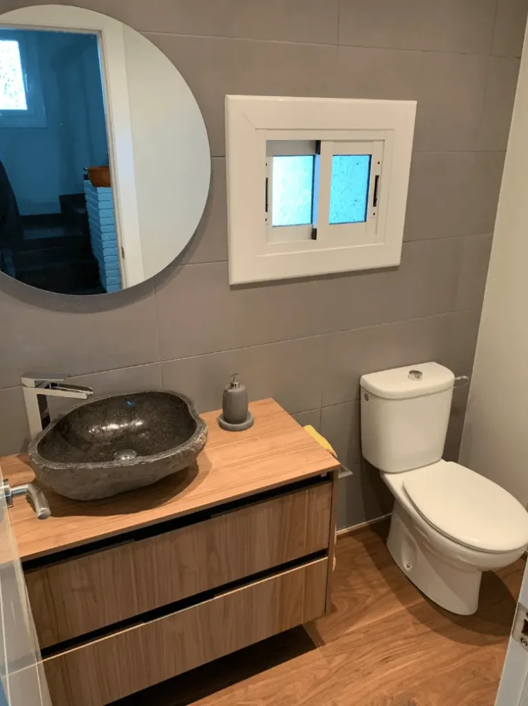 Lavamanos de piedra natural de forma orgánica sobre encimera de madera clara, con grifería cromada y espejo redondo en baño moderno.