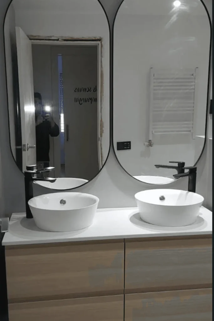 Lavamanos doble de cerámica blanca sobre encimera blanca con grifería negra, mueble de madera y espejos verticales de marco negro.