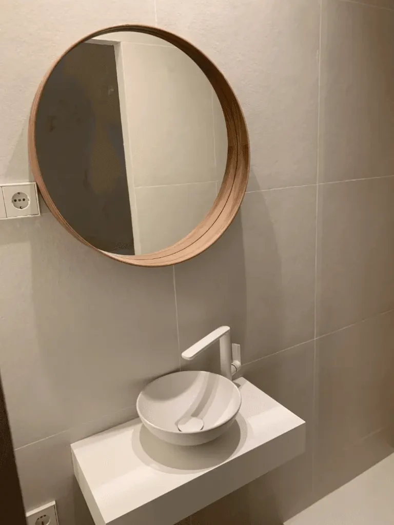 Lavamanos redondo blanco sobre encimera flotante blanca con grifería moderna y espejo redondo de madera en baño minimalista.