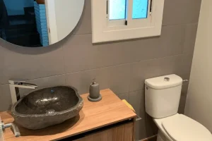 Lavamanos de piedra natural de forma orgánica sobre encimera de madera clara, con grifería cromada y espejo redondo en baño moderno.