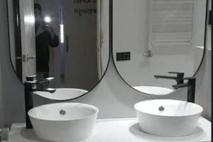 Lavamanos doble de cerámica blanca sobre encimera blanca con grifería negra, mueble de madera y espejos verticales de marco negro.