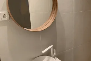 Lavamanos redondo blanco sobre encimera flotante blanca con grifería moderna y espejo redondo de madera en baño minimalista.