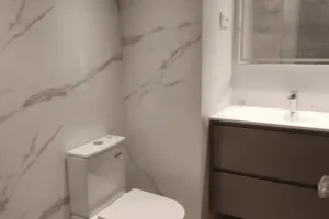 Baño moderno con sanitario de líneas rectas, lavabo suspendido y revestimiento de mármol blanco veteado.